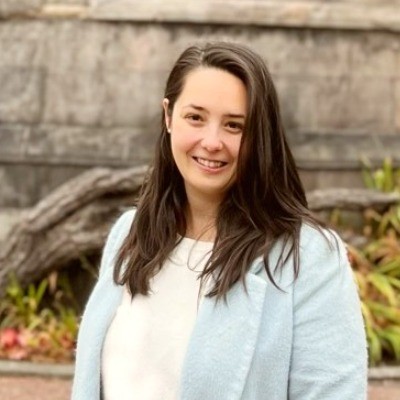 The image depicts Lili, a woman with brown hair, brown eyes, and fair skin. Lili is wearing a light blue coat over a white jumper. She is standing in front of a blurred background featuring some greenery and granite blocks of an old building.